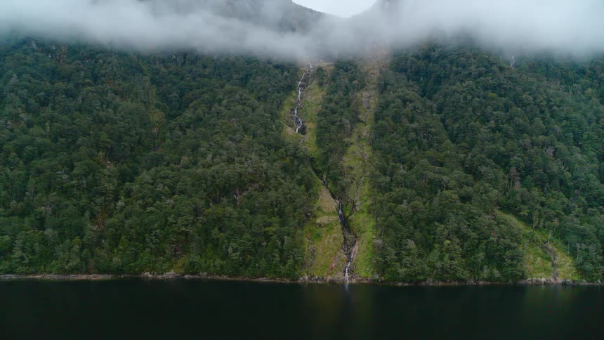 Waterfall flows down slope through forest covered terrain under low lying clouds as powered boat speeds past
