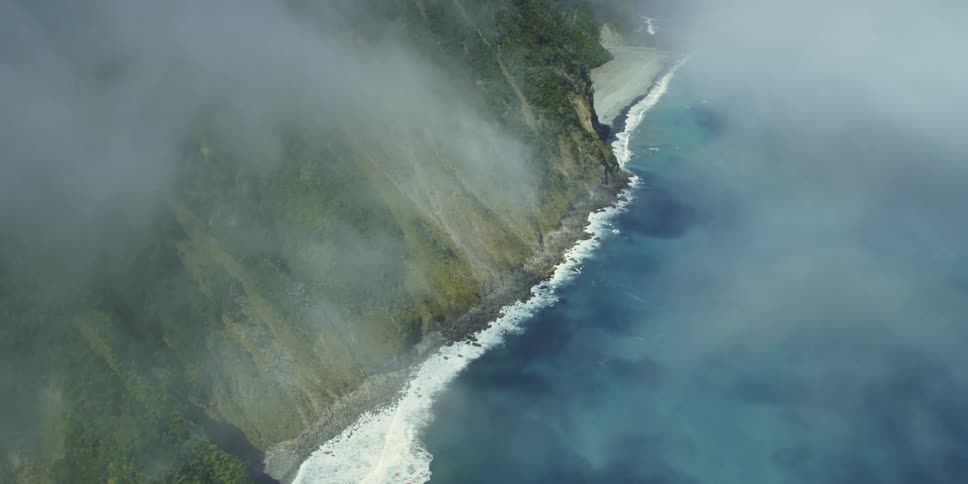Wispy clouds hang over the blue ocean waves and foam along the cliffside shore