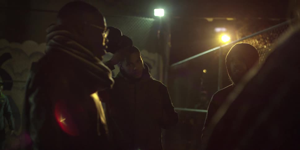 A group of men stand outside at night with street lights and a chain link fence in the background