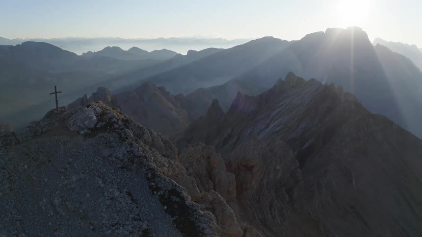 Sun shines over peak in mountain covered in pockets of fog including lone cross on mountain