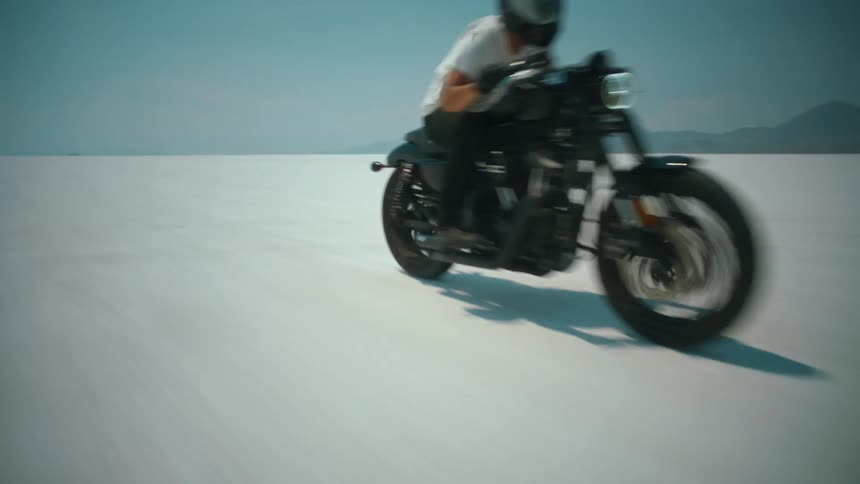 A person drives a motorcycle across flat terrain with a mountain range in the distance