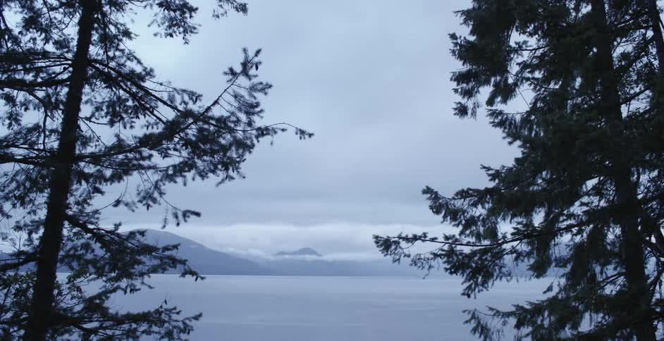 A time lapse shows clouds passing above a large body of water lined with mountains and evergreen trees