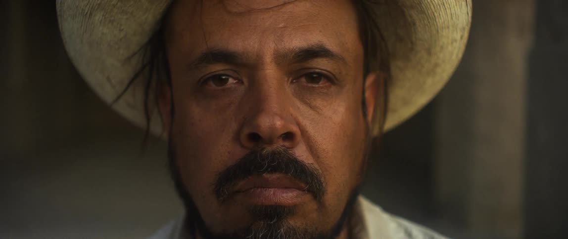 A man with a hat and shirt in a room standing and staring with a wall in the background
