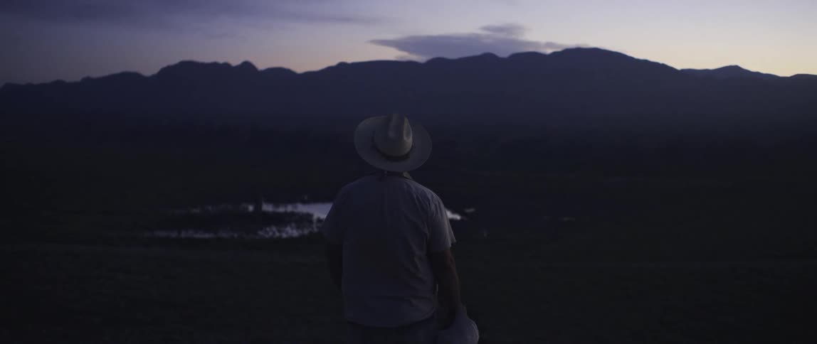 A man with a hat standing with a landscape with a pond a mountain range the sky and clouds in the background