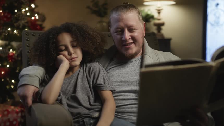 A fathers sits with his sleepy daughter while reading a book to her