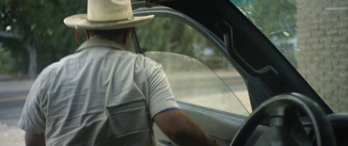 Man with beard brown hat and shortsleeved shirt sits in car looking up at building and gets out of car