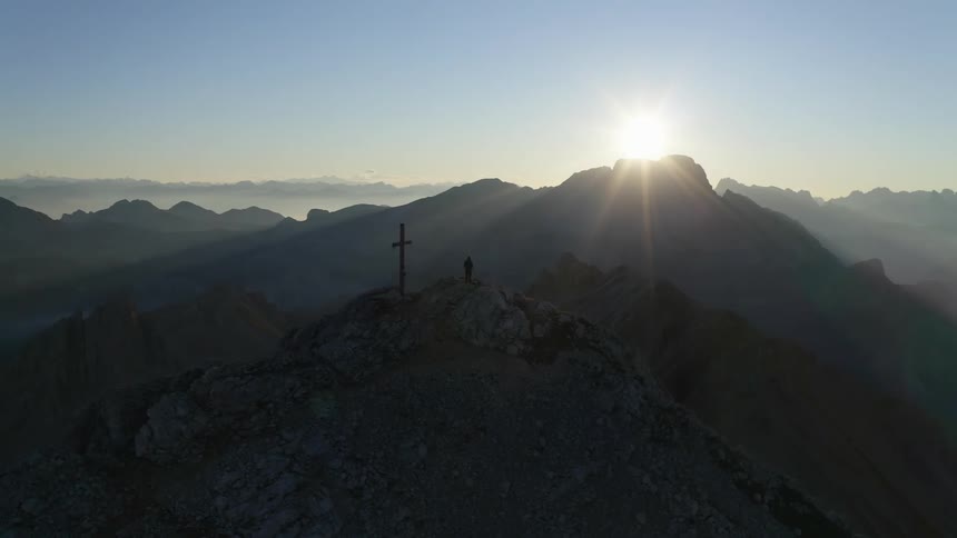 Sunlight shines over a distant peak onto a misty mountain range where a cross and statue have been erected