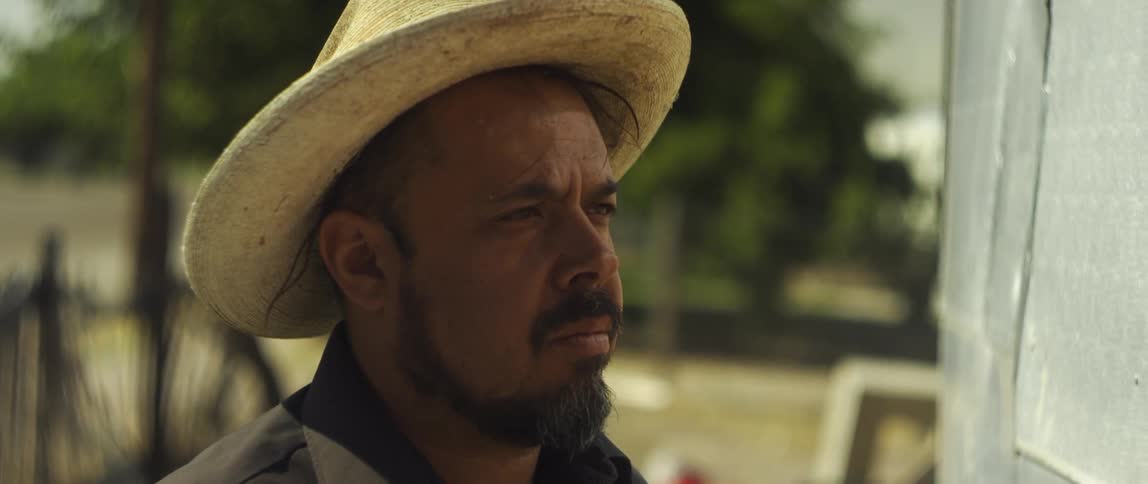 A man with a hat and a shirt standing with a fence and trees in the background