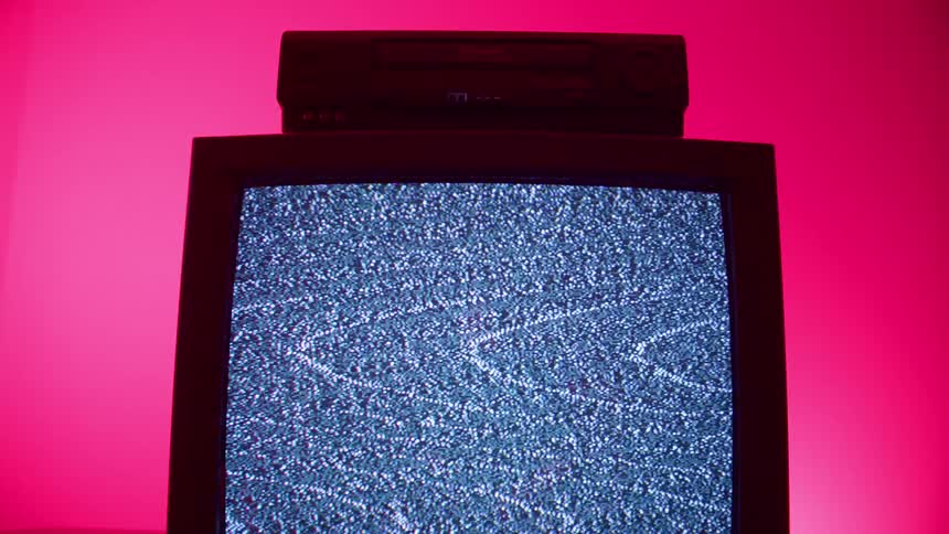 A crt television with a video cassette player resting on top displays static in a pink illuminated room