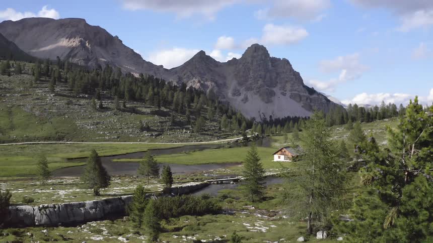 Cabin sits on field near winding river in valley leading to craggy mountain under cloudy sky