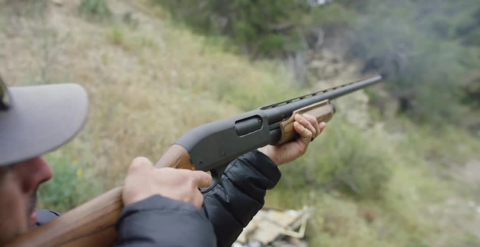 A man shoots a rifle near tall grass covered hills