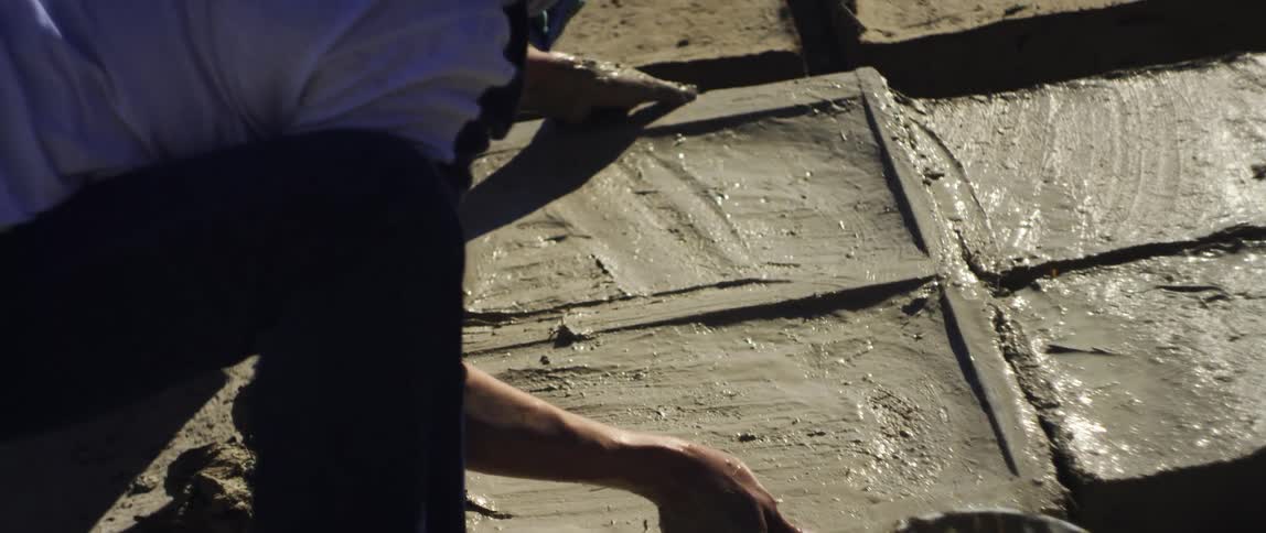 A person wets their hands before carefully lifting a wooden frame from around wet clay being shaped into rectangular tiles