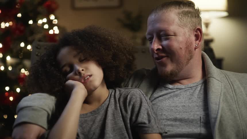 Father with earring and grey jacket kisses daughter with curly hair sleeping in front of christmas tree