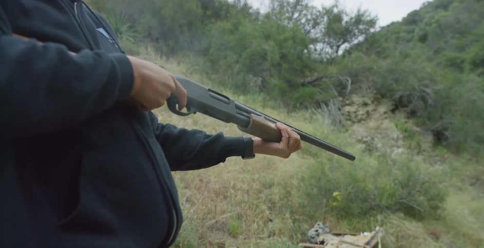 A man with a hat and sunglasses shooting a rifle with trees the sky and clouds in the background
