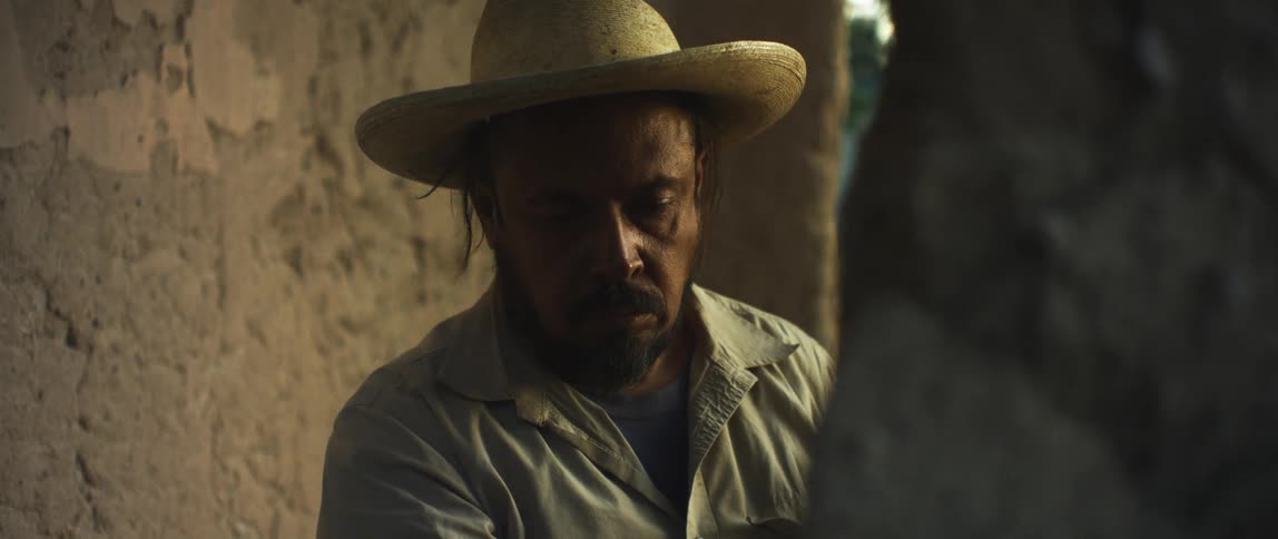 Man wearing straw hat concentrates downward standing near adobe wall across street from one story house