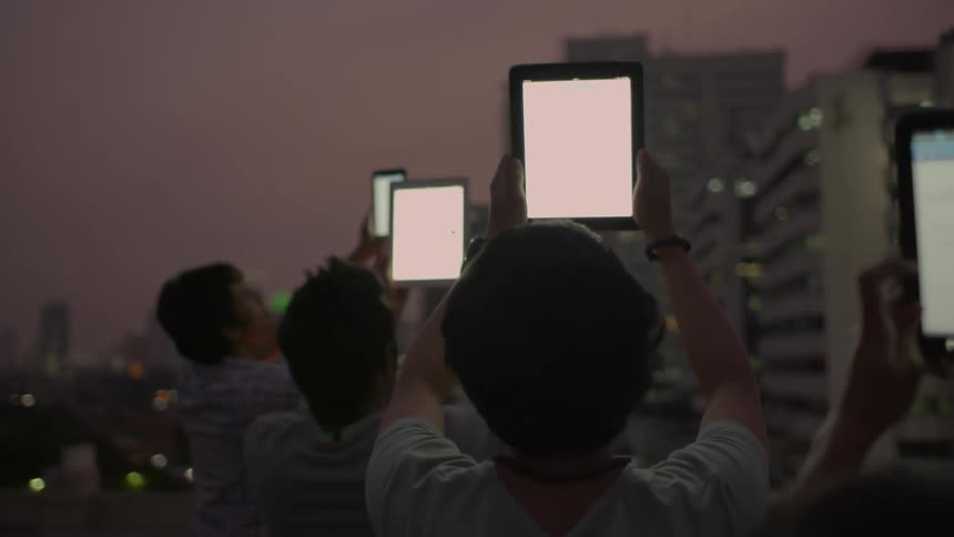 Young boys hold up their tablets as they stand along the edge of a rooftop downtown