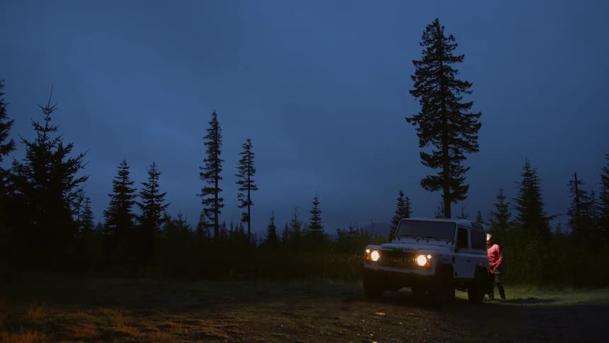 A man walks to the back of a land rover in the rain then gets back in