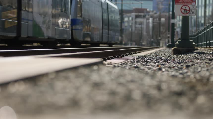 Light rail moves along tracks away from intersection as car crosses tracks with buildings in the background