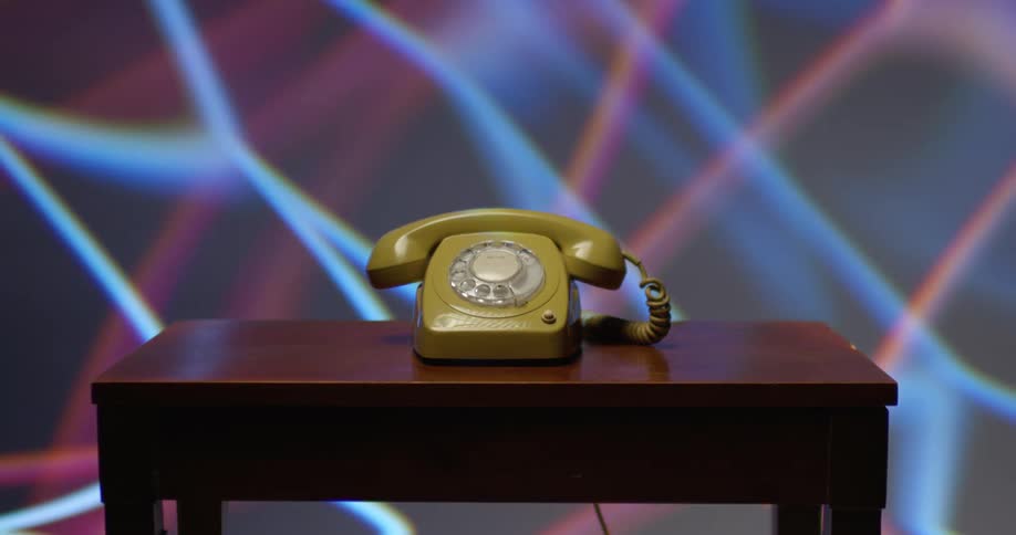 A yellow rotary telephone is on a table with colored lights moving on the wall behind it