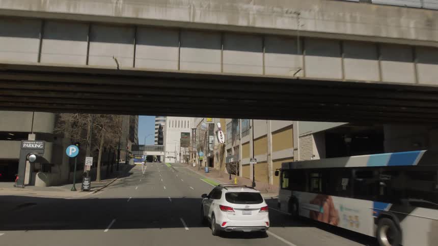 A white automobile drives along a city street while passing beneath an overpass