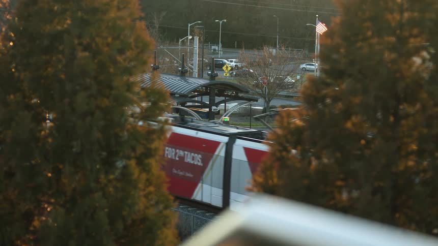 A train running on a train track with trees in the foreground with an american flag streetlights and vehicles in the background