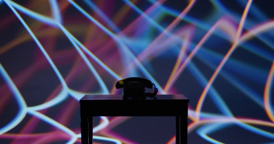 An older rotary dial phone is atop a table in a dark room with light projections displaying colored lines