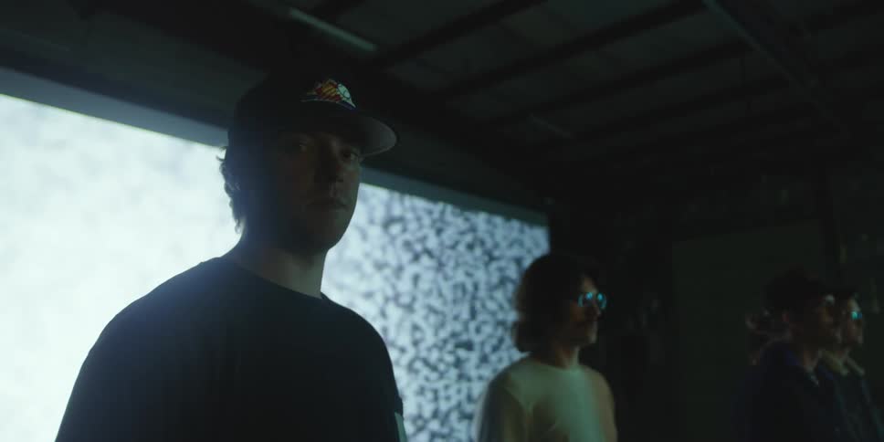 A man standing with a group of men in front of a large screen in a dimly lit room stares into the camera