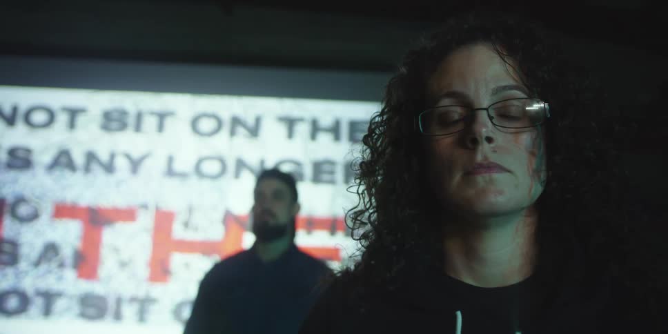 Woman with glasses and curly hair and man with beard stand in front of giant screen protesting