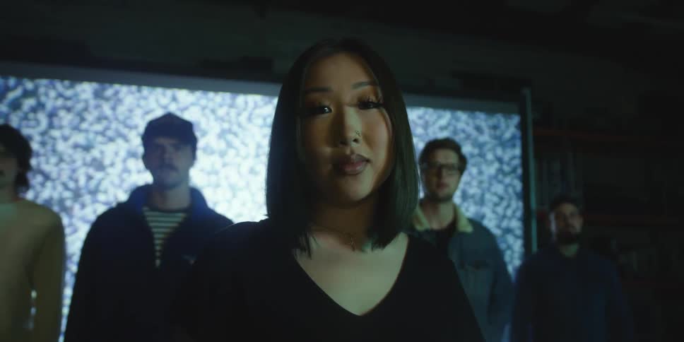 A woman stares into the camera while standing in front of a line of men who are in turn standing in front of a large video wall