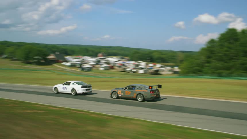 Race cars speed along race track bordered by woods under a cloudy blue sky