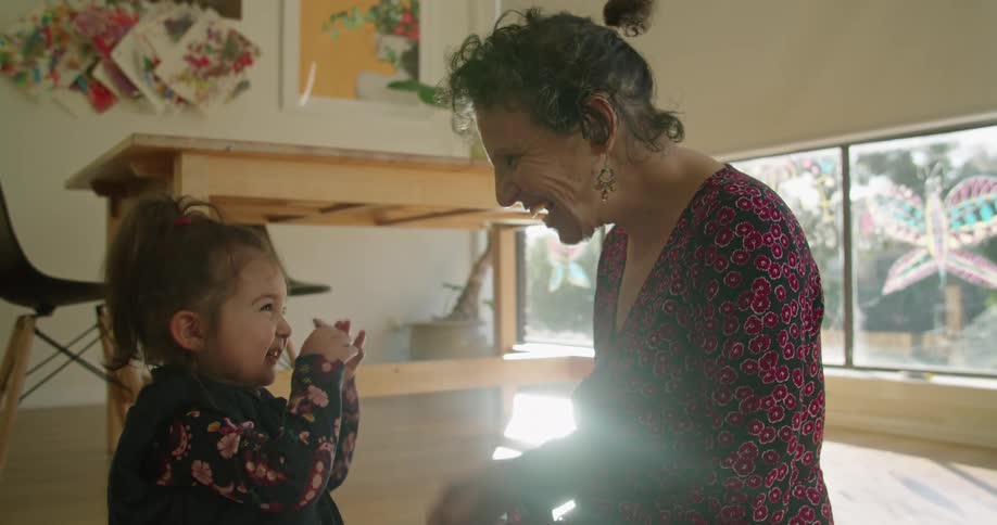 A woman kneels on the floor and gives something to a young girl who gets very excited for the item