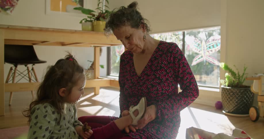 An elderly woman sits on the floor in a home andputs shoes on a little girl