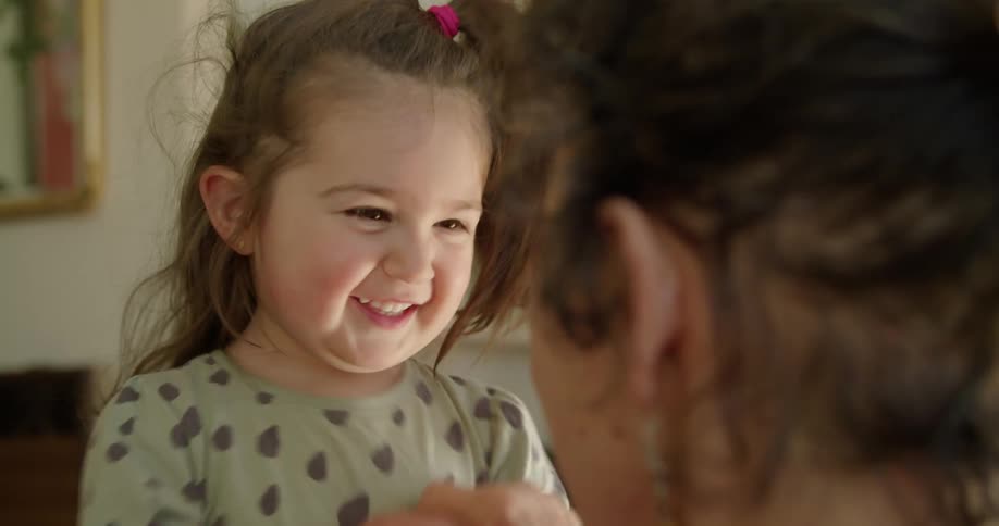 A woman plays with the hands of a smiling young girl in a house during the day