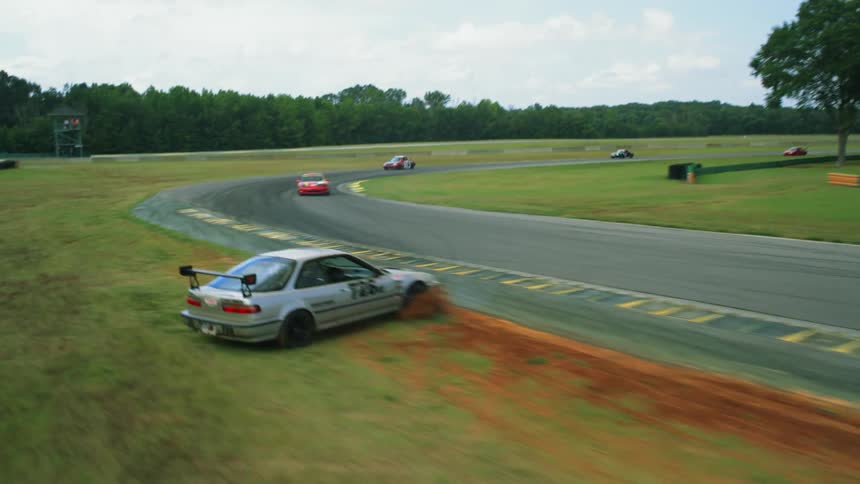 Multiple vehicles drive around a race track and one slides off to the side grass