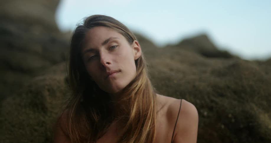 A woman uses her hand to tuck her hair behind her ear as she sits outdoors on a rock formation while looking into the camera