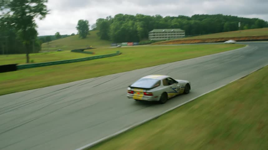 Cars race on a racetrack under a cloudy sky as a forest and a building sit in the distance