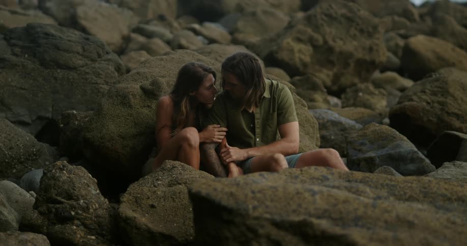 A woman holds onto her boyfriends arm and they kiss while sitting against rocks along the coast