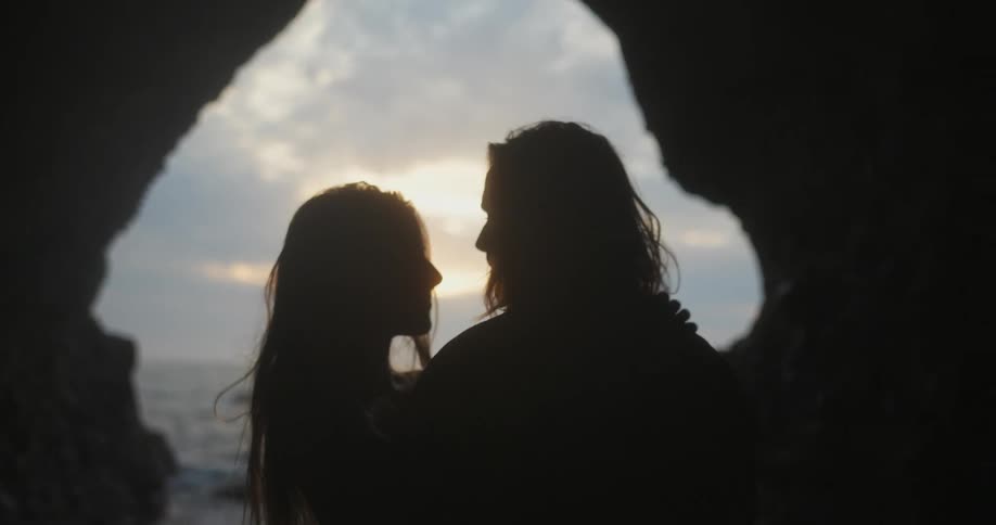 A couple in silhouette stand just inside a cave near a body of water