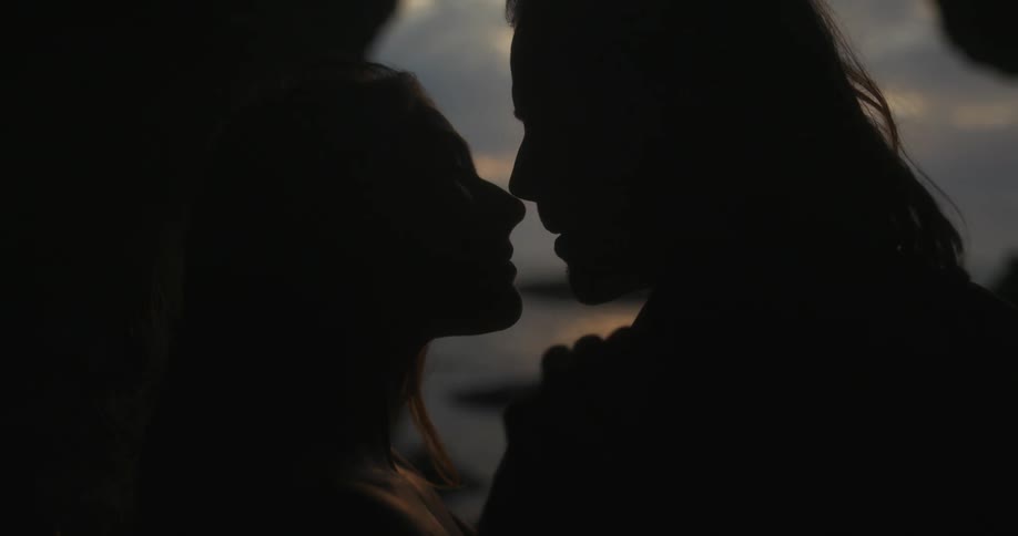 Man with long hair and woman with brown hair kiss each other and are silhouetted against the sky in cave at dusk