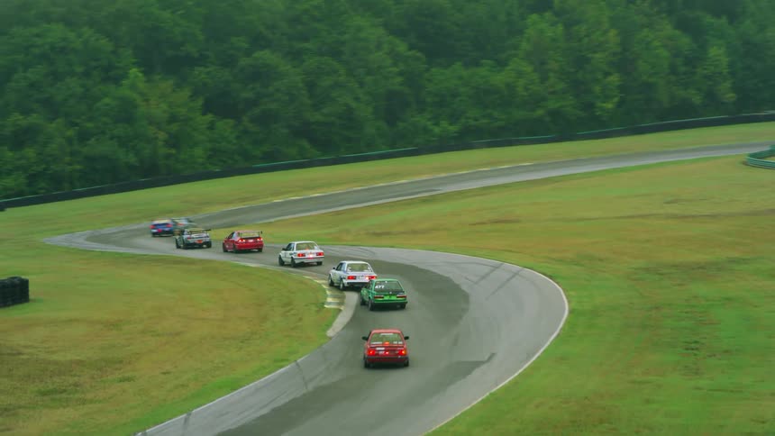 A racetrack surrounded by grass with cars racing on the track with trees in the background