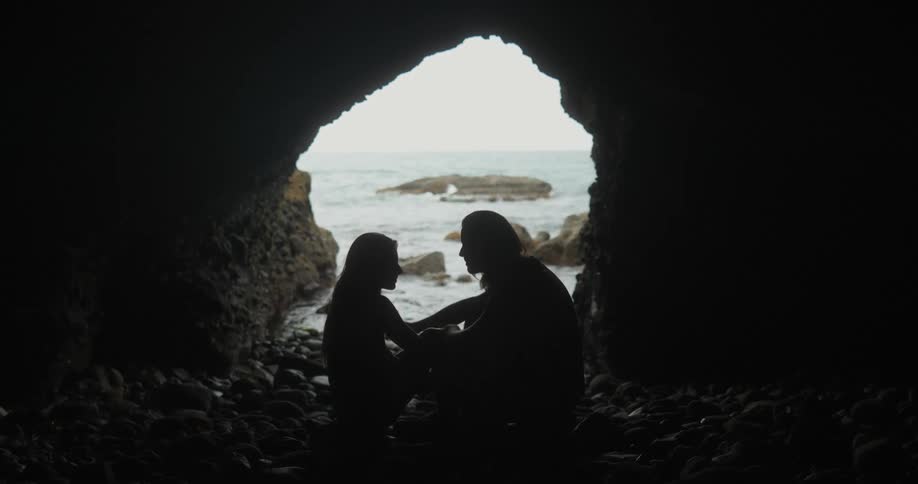 Couple sit near mouth of sea cave on rocks talking with man touching woman leg