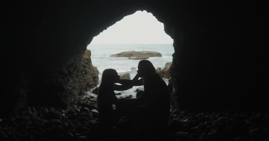 A man and woman sit on rocks in a cave caressing each other while waves splash at the entrance