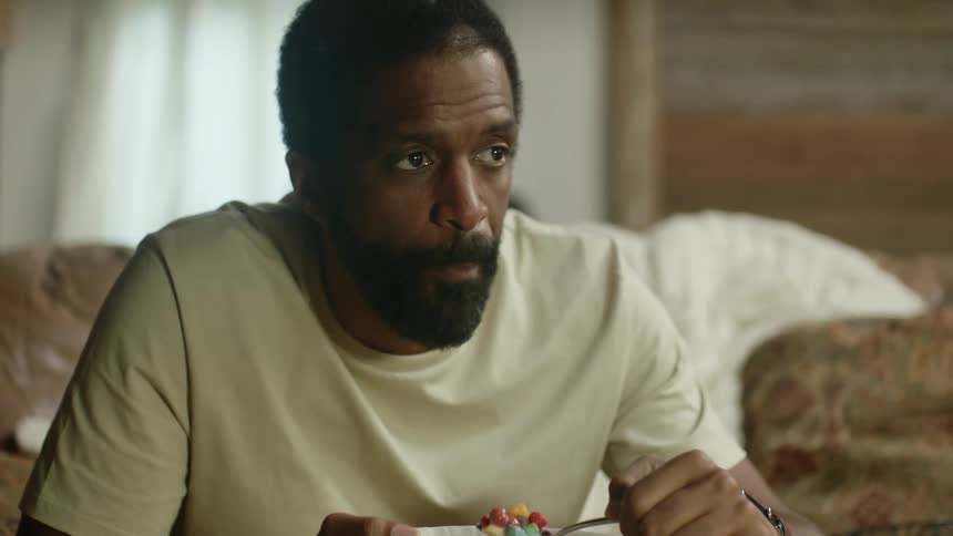 A man sits on a couch eating from a bowl and laughs to himself in amusement