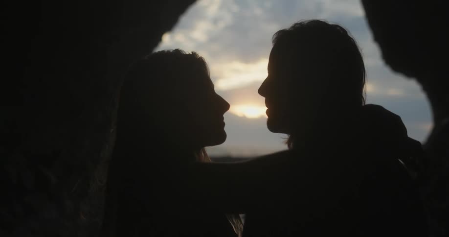 Couple hug and kiss backlit by light coming from pink tinted sky over ocean and through cave mouth