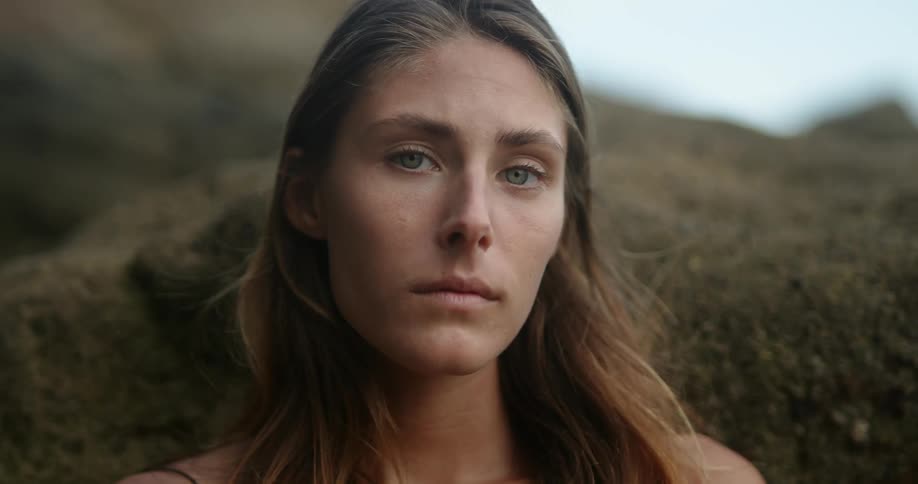 Woman with brown hair faces the camera in front of beach rock by the sea