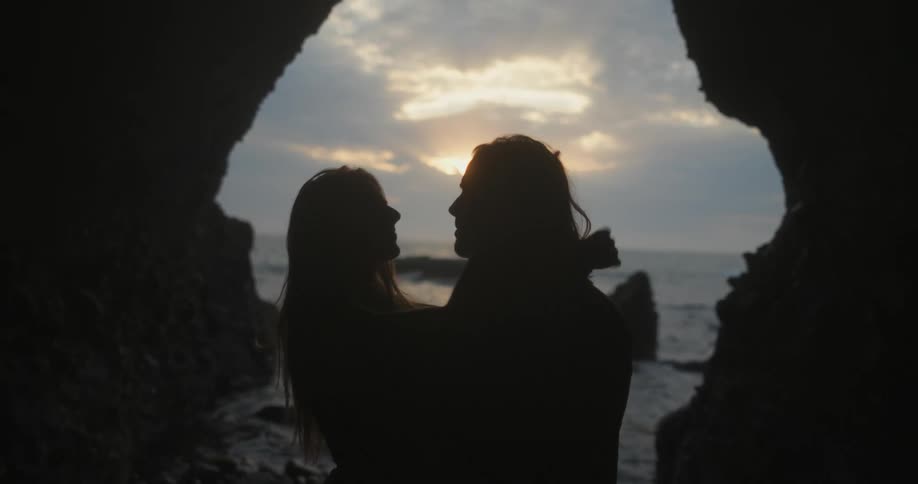 A man and woman stand close to each other romantically standing in a cave by the ocean