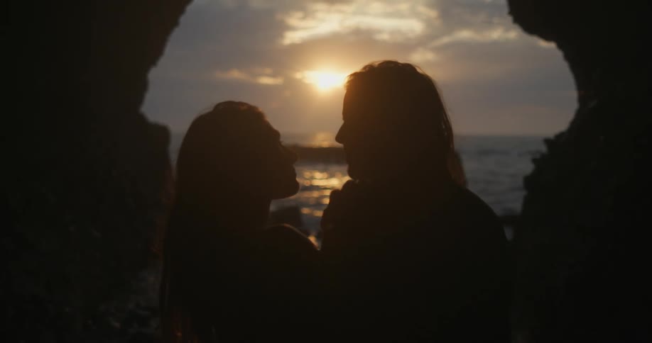A man and woman stand on the shore and look in to each others eyes as the sun sets