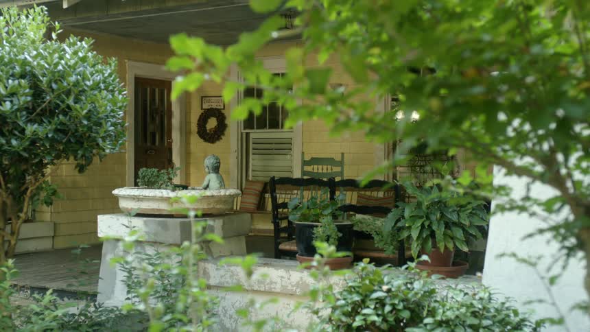 Statue sits in rectangular plant pot on top of porch decorated with wicker love seat and chair