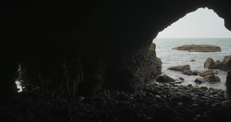 Two people wearing shorts walk over stones in dark cave with mouth showing ocean and horizon