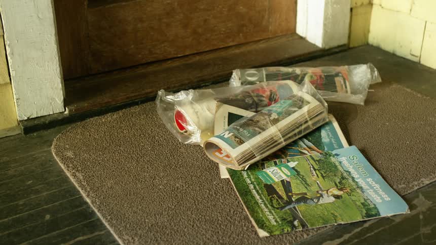 A magazine falls onto a stack of magazines and newspaper in front of the door of a house